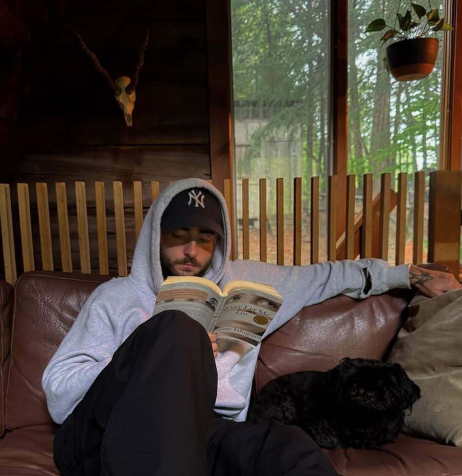 A man reading a book on a sofa beside his dog